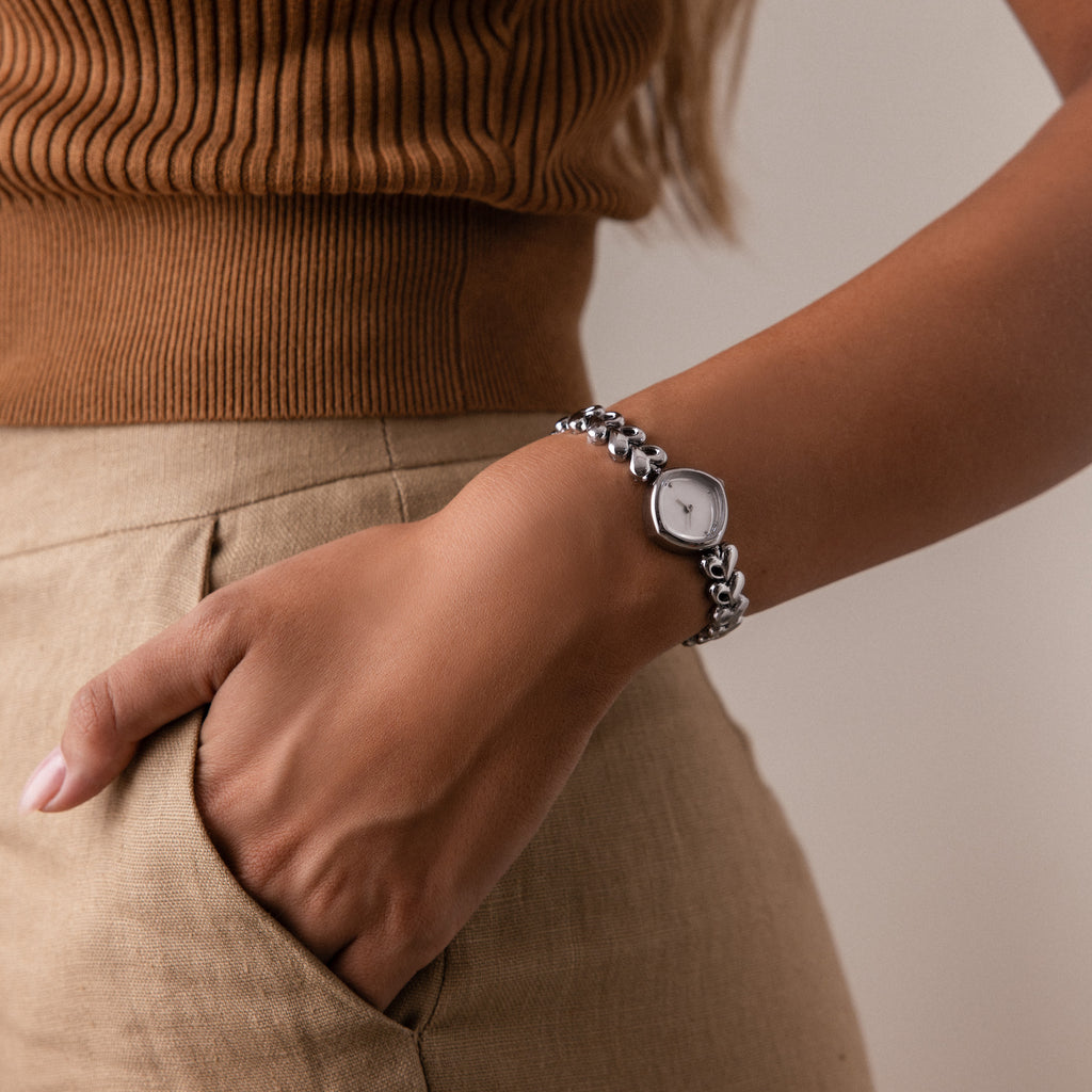 A woman dressed in brown stands with her hand in her pocket, wearing the Heart Link Timepiece in White Gold, featuring a minimalist dial.