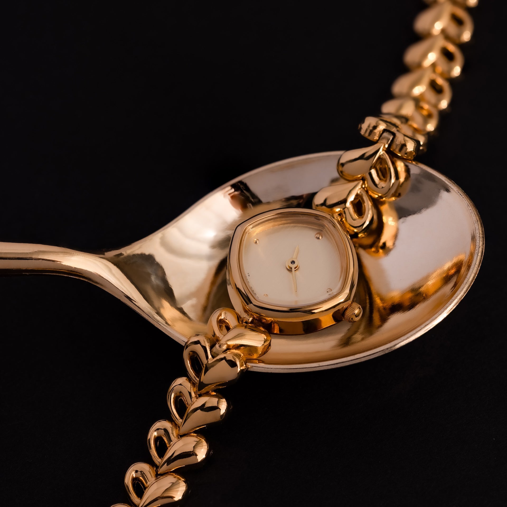 The Heart Link Timepiece in 18K Gold, featuring heart-shaped links and a minimalist dial, rests on a shiny gold spoon against a black background.