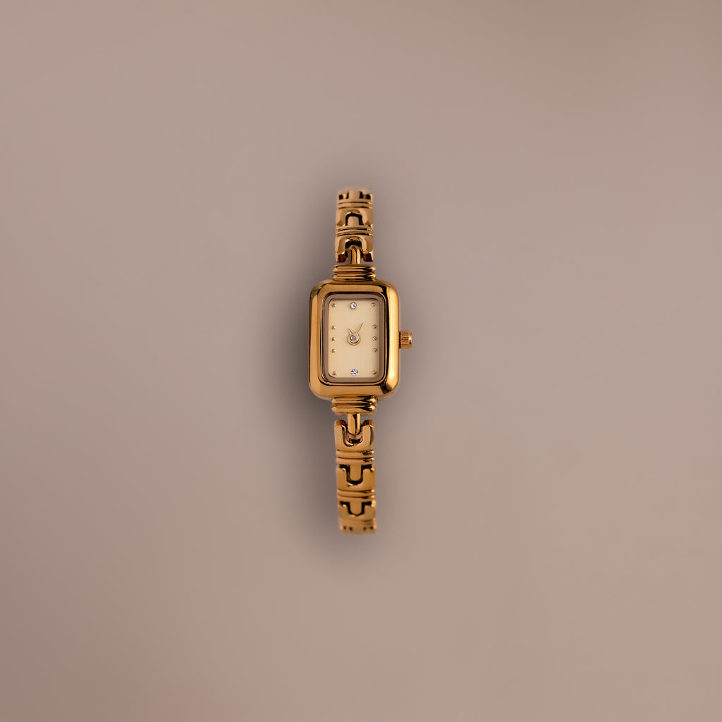 The Azteca Link Timepiece in 18K Gold features a rectangular case and minimalist champagne dial, with a patterned gold band, displayed elegantly on a plain light brown background.