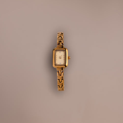 The Azteca Link Timepiece in 18K Gold features a rectangular case and minimalist champagne dial, with a patterned gold band, displayed elegantly on a plain light brown background.