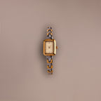 The Azteca Link Timepiece in Mixed Metal, featuring a rectangular case, patterned band, and minimalist champagne dial, rests on a plain beige background.