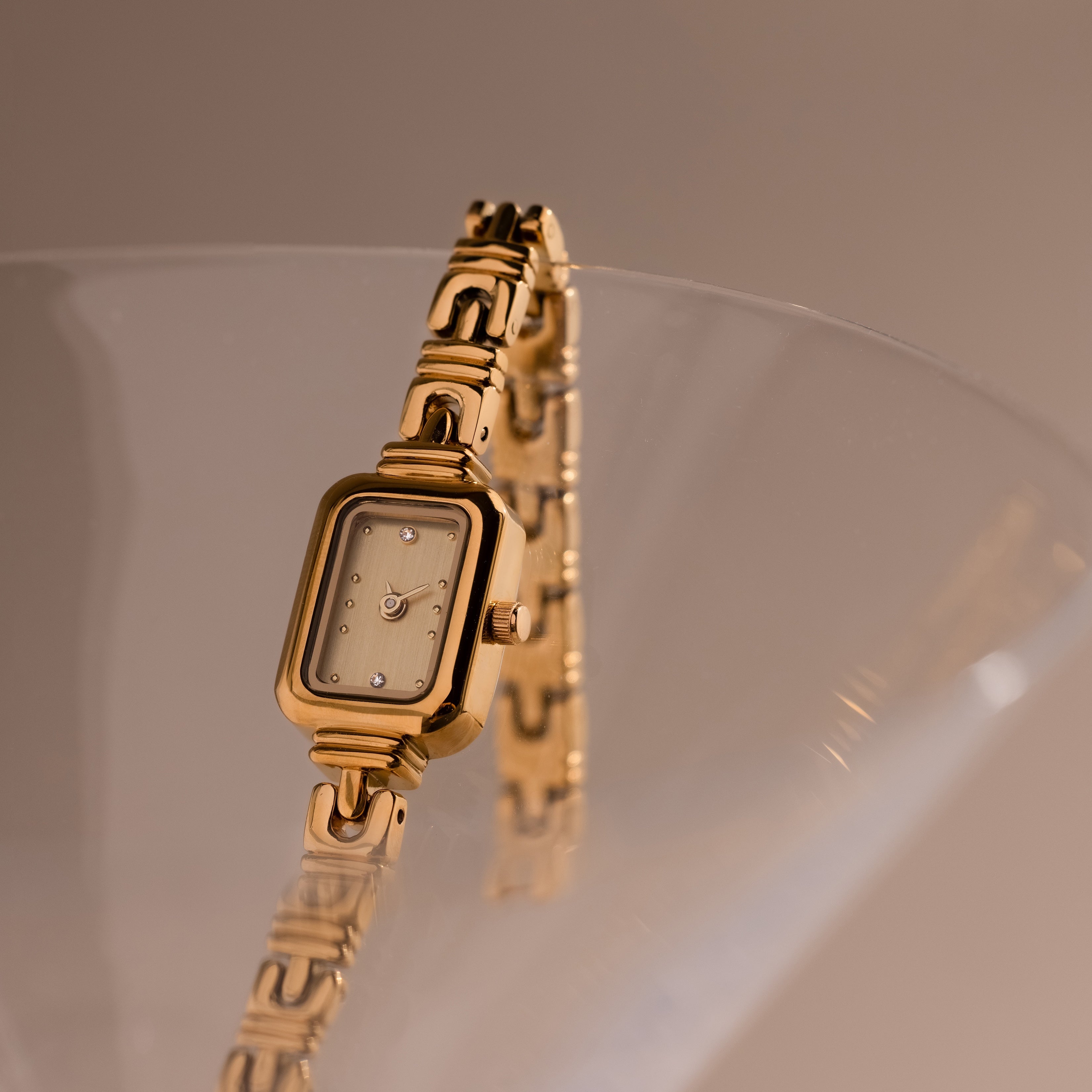 The Azteca Link Timepiece in 18K Gold, featuring a minimalist champagne dial and rectangular case, is elegantly displayed on the rim of a clear glass.