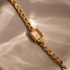 Azteca Link Timepiece with a rectangular case and minimalist champagne dial sits on a textured beige surface, its face illuminated by soft, wavy light patterns.