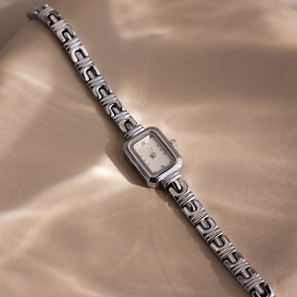 Azteca Link Timepiece in White Gold, featuring a minimalist champagne dial and interlocking bracelet, rests on a beige surface in soft, dappled sunlight.