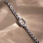 Azteca Link Timepiece in White Gold, featuring a minimalist champagne dial and interlocking bracelet, rests on a beige surface in soft, dappled sunlight.