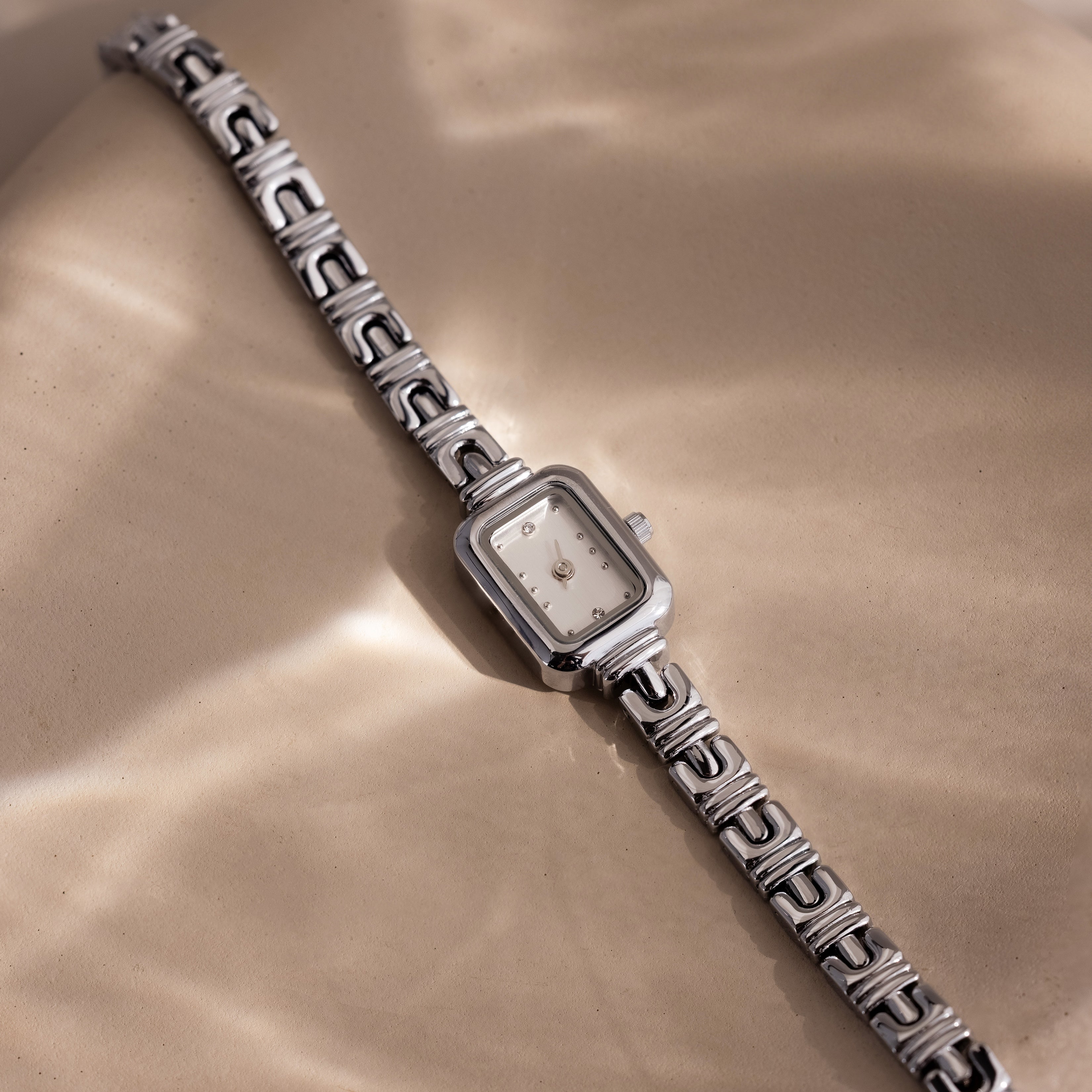 Azteca Link Timepiece in White Gold, featuring a minimalist champagne dial and interlocking bracelet, rests on a beige surface in soft, dappled sunlight.