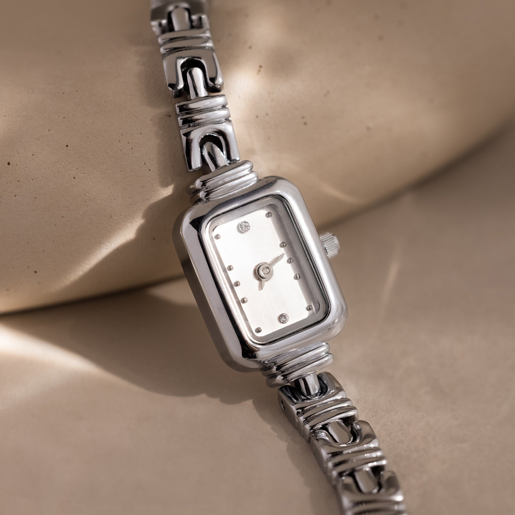 The Azteca Link Timepiece, with its rectangular case and patterned metal band in silver, is displayed on a beige surface.