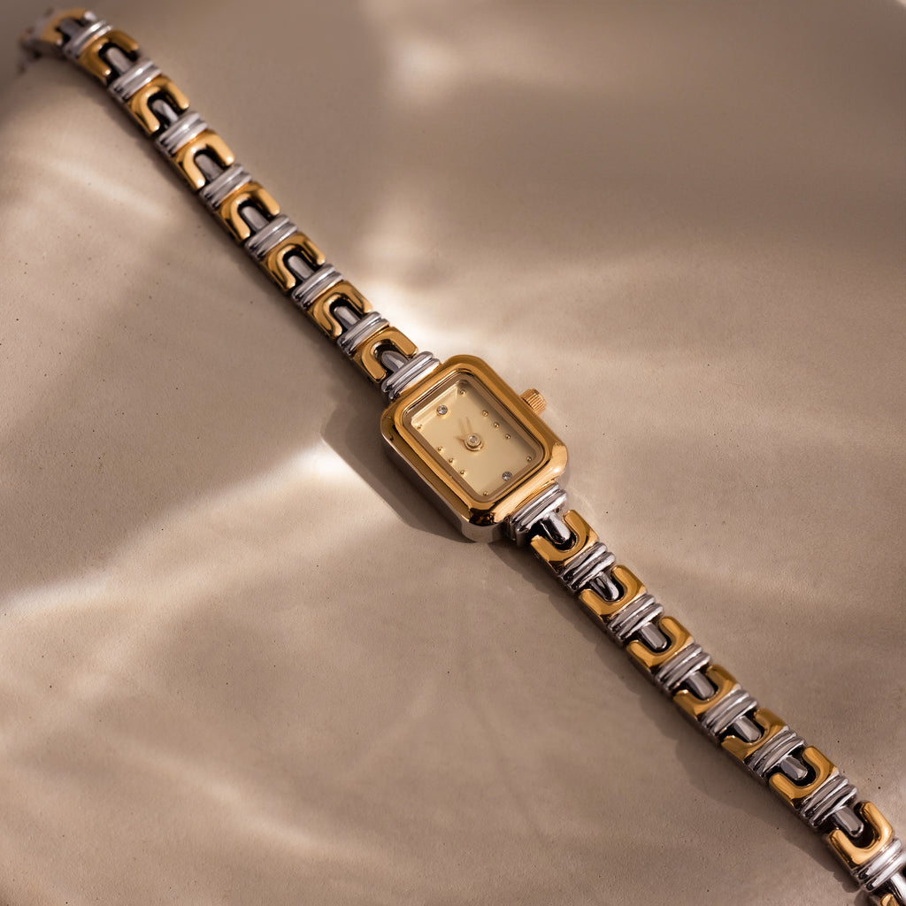 The Azteca Link Timepiece in Mixed Metal, featuring a rectangular case, rests on a beige surface, reflecting soft, wavy light.