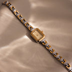 The Azteca Link Timepiece in Mixed Metal, featuring a rectangular case, rests on a beige surface, reflecting soft, wavy light.