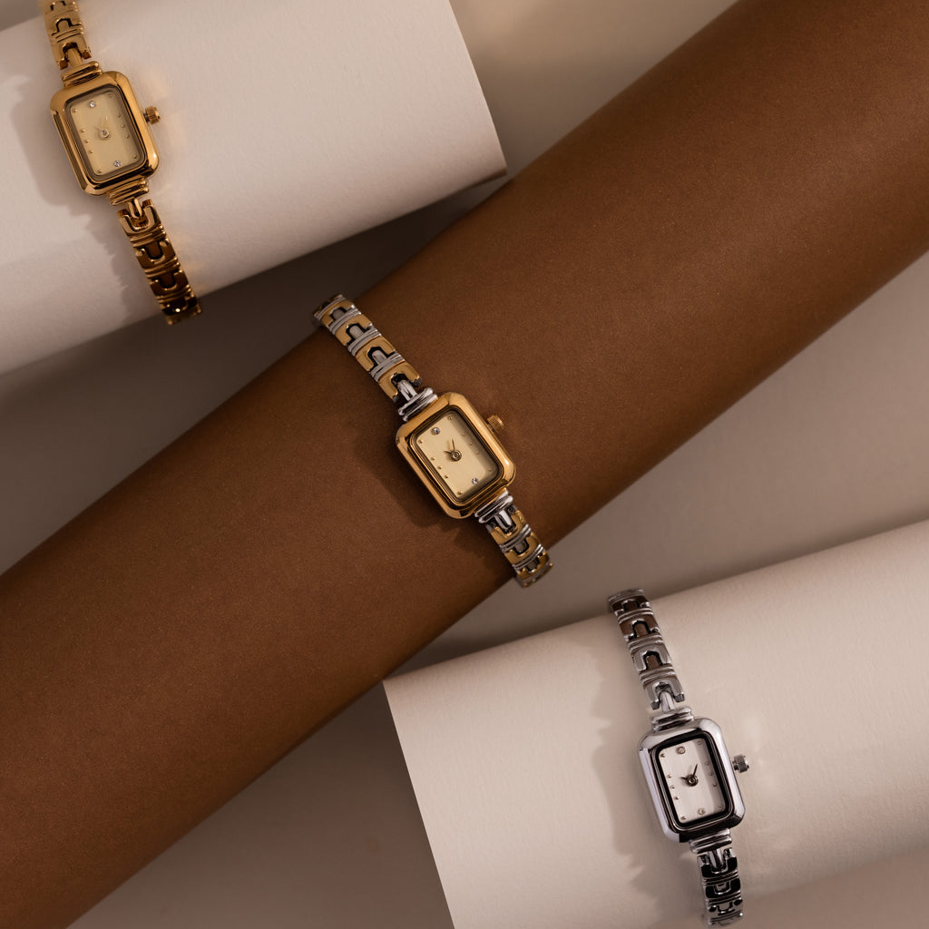 Three elegant wristwatches, including the Azteca Link Timepiece and a minimalist champagne dial model, are displayed in gold and silver on brown and white cylindrical stands.