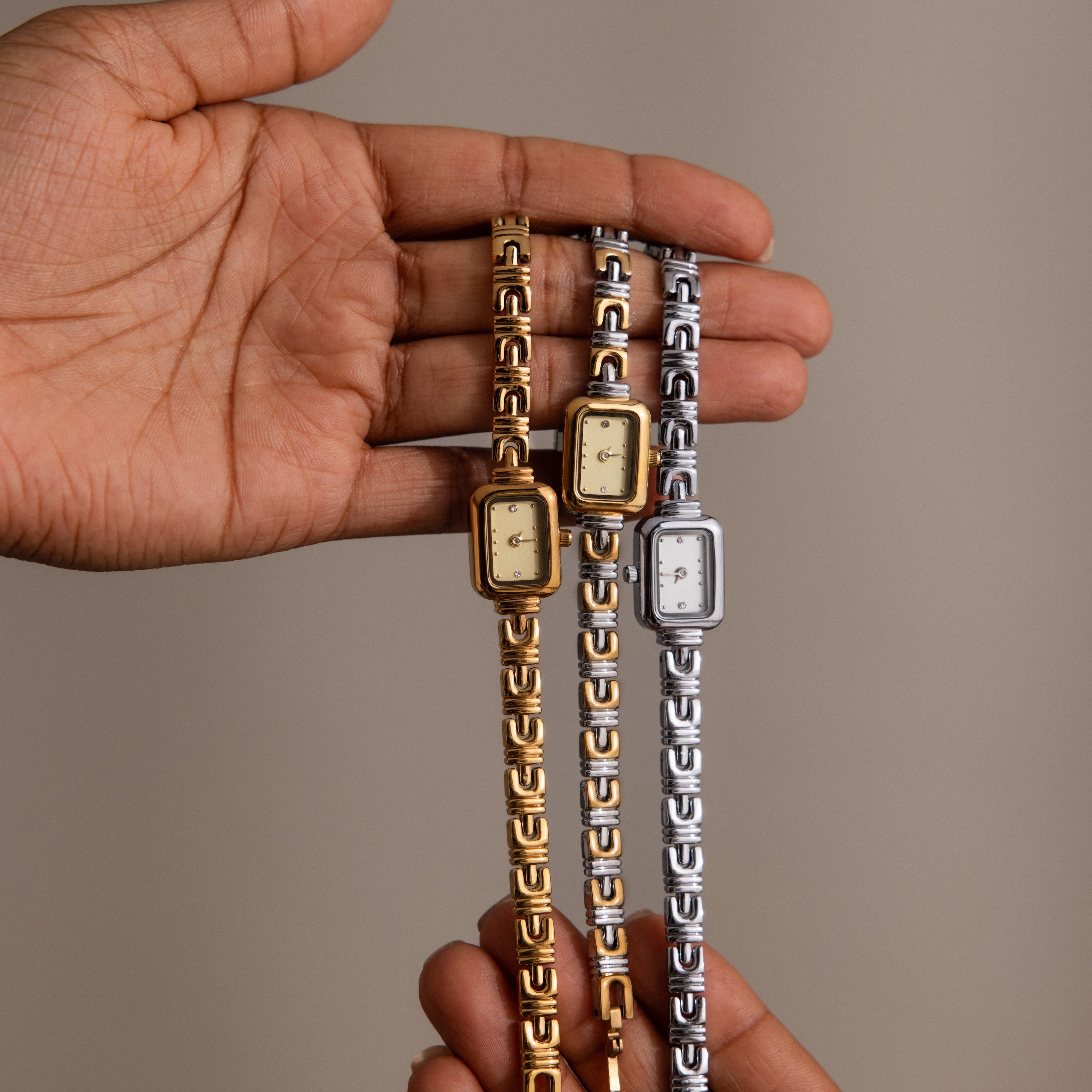 A hand holding two gold and one silver rectangular case watches with patterned metal bands, featuring the Azteca Link Timepiece and a minimalist champagne dial.