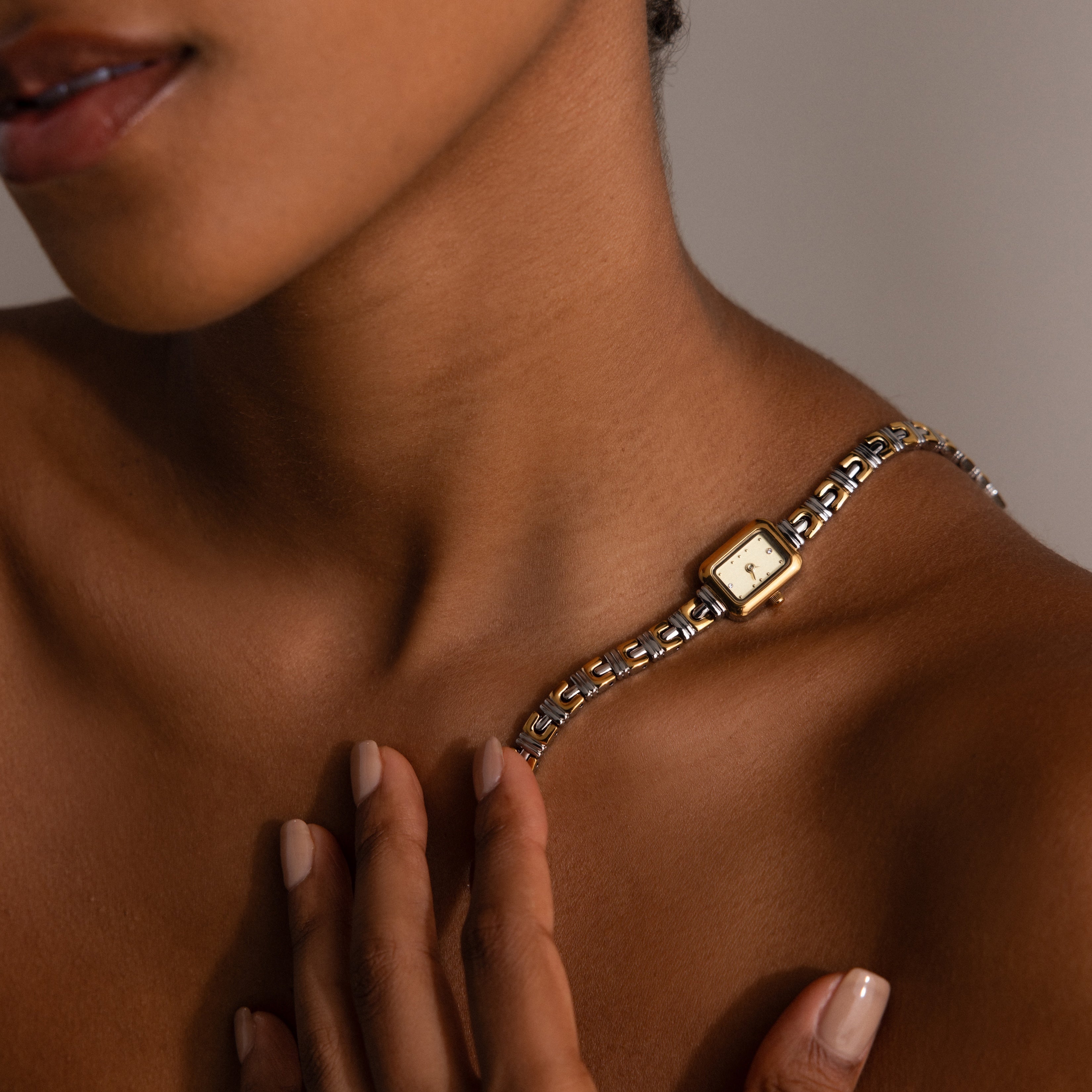 The Azteca Link Timepiece in Mixed Metal features a minimalist champagne dial, showcased on a gold wristwatch resting elegantly against a woman's bare shoulder as her hand traces her collarbone.