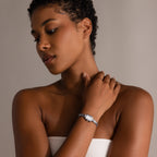 Woman with short curly hair, wearing a strapless top and an Azteca Link Timepiece in White Gold with a minimalist champagne dial, looks down thoughtfully.