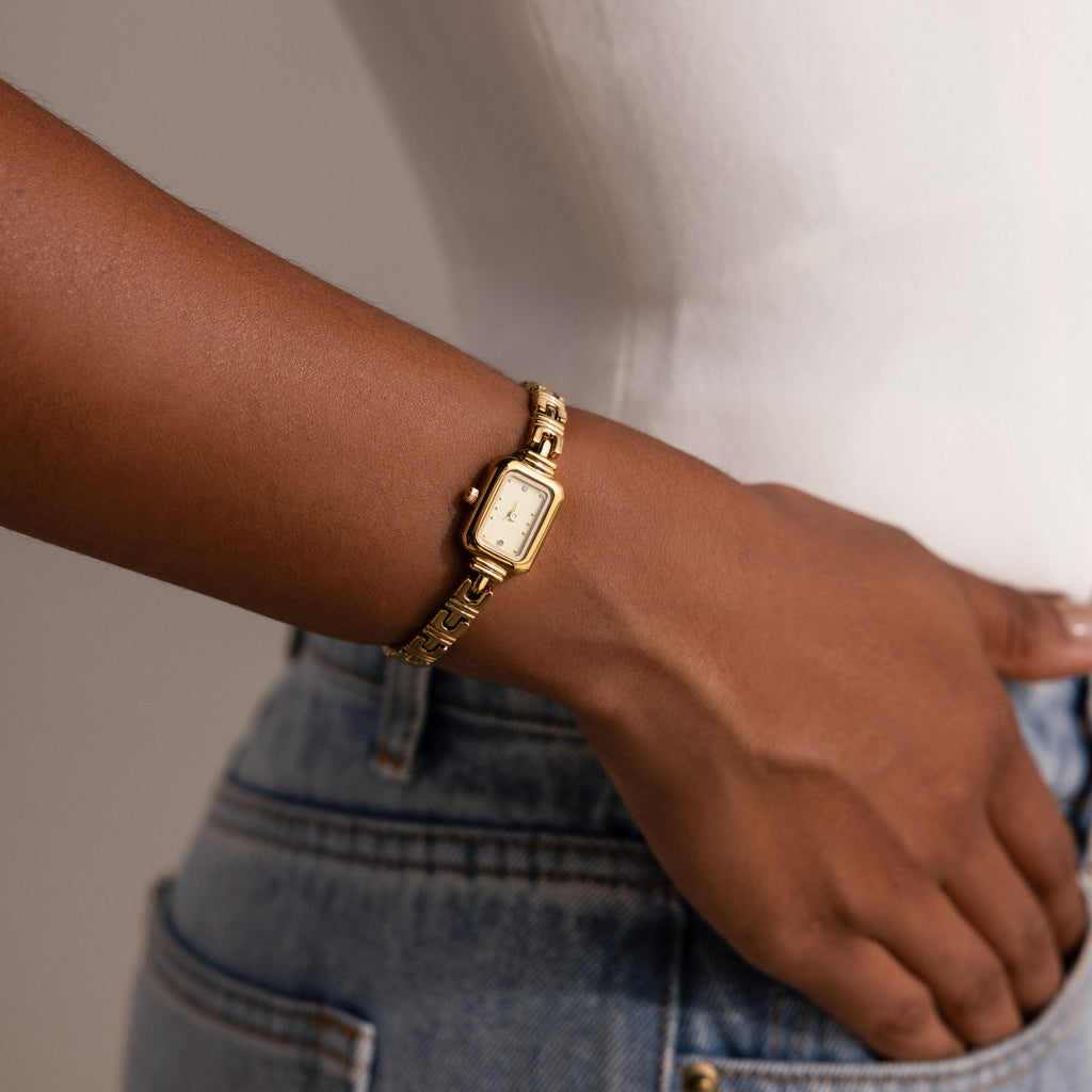 An Azteca Link Timepiece in 18K Gold with a minimalist champagne dial adorns a woman's wrist, styled with blue jeans and a white top.