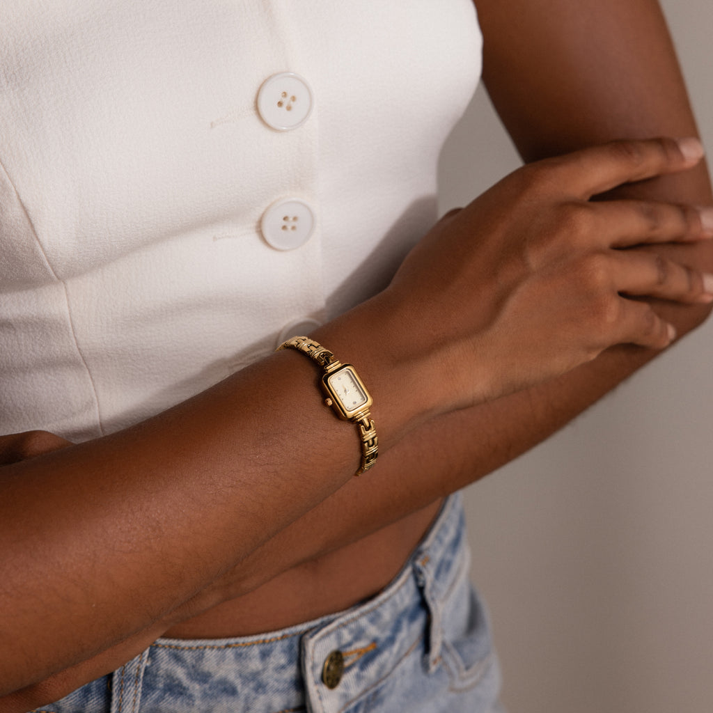 A woman in a white top and jeans crosses her arms, wearing the Azteca Link Timepiece in 18K Gold with a minimalist champagne dial.
