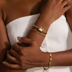 A woman in a white strapless top wears the Azteca Link Timepiece in 18K Gold with a minimalist champagne dial and gold bracelets, her arms crossed confidently.