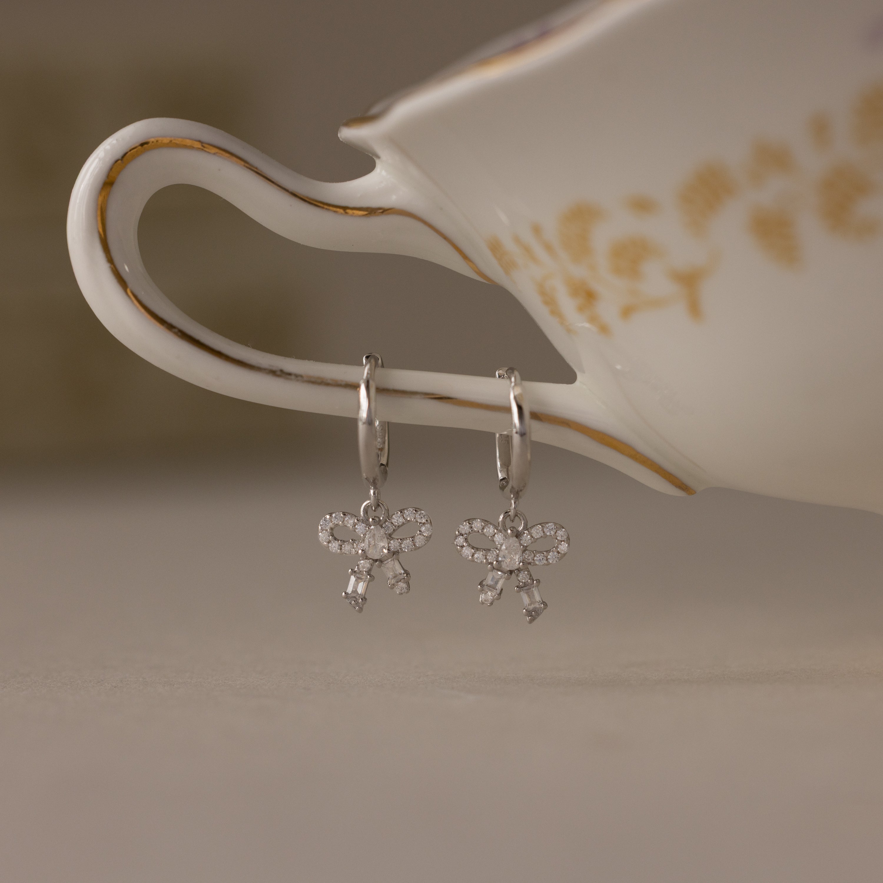 Pave Bow Charm Hoops earrings in Silver hanging from a teacup with floral patterns.