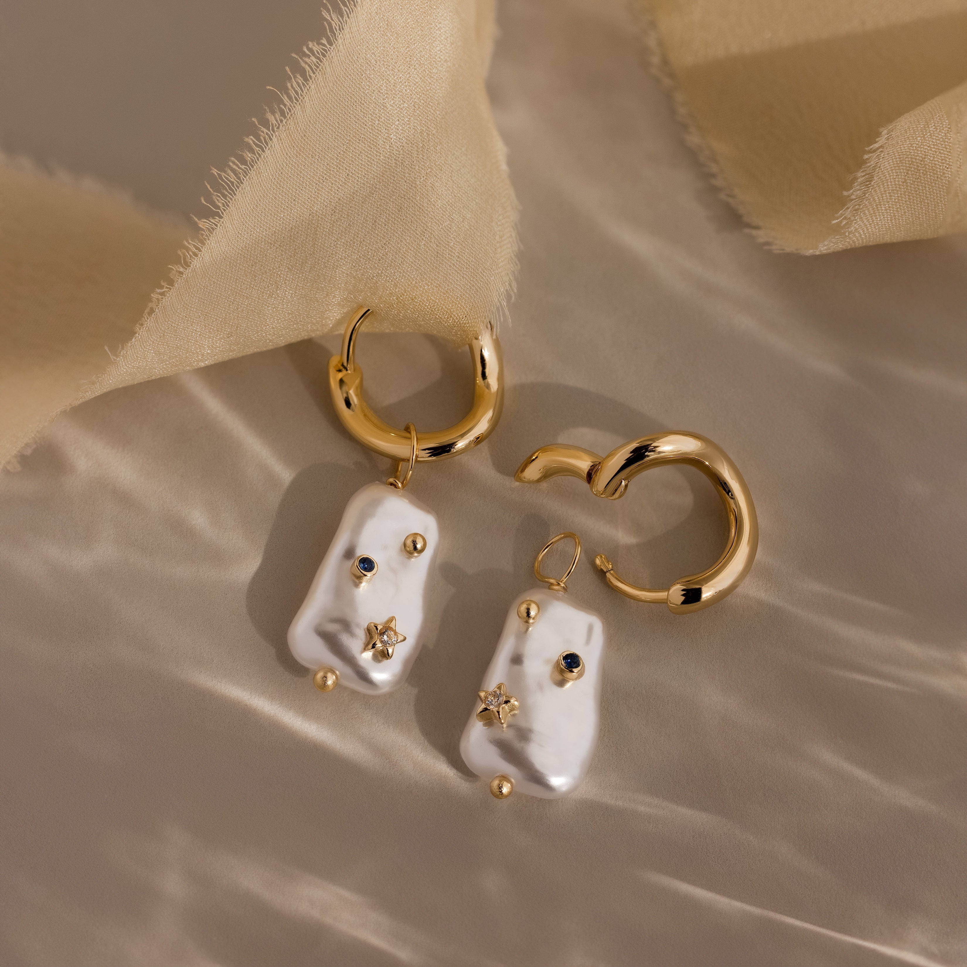 The Starry Baroque Pearl Charm Hoops feature irregular pearls, tiny gold stars, and sparkling gems—Baroque-style earrings beautifully displayed on soft beige fabric.