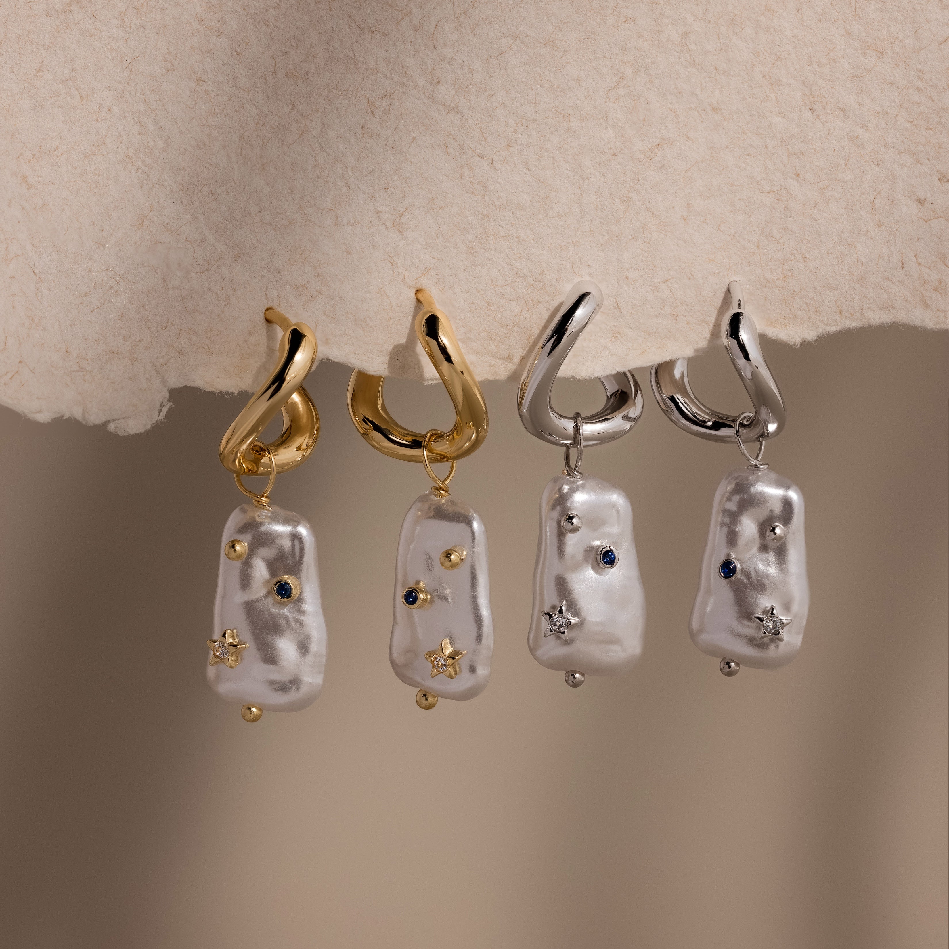The Starry Baroque Pearl Charm Hoops in 18K Gold, featuring irregular Baroque pearls and gemstone charms, are displayed on a beige background.