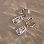 The Starry Baroque Pearl Charm Hoops in White Gold feature irregular pearl-like charms, blue stones, and star accents, capturing a unique allure reminiscent of Baroque pearl earrings, shown on a beige surface.