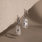 Starry Baroque Pearl Charm Hoops in White Gold feature irregular pearl drops with blue and silver accents, displayed on a textured beige surface.