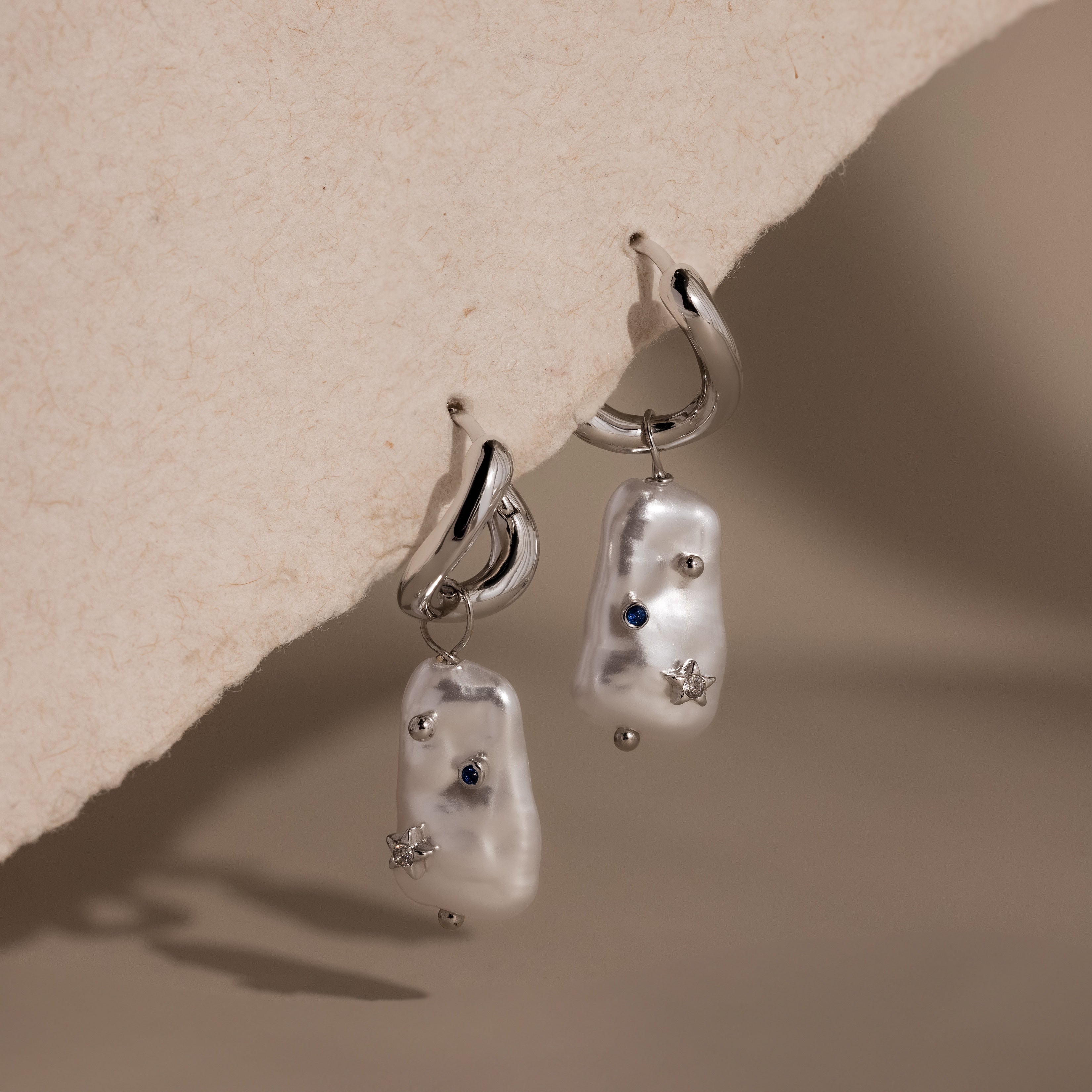 Starry Baroque Pearl Charm Hoops in White Gold feature irregular pearl drops with blue and silver accents, displayed on a textured beige surface.
