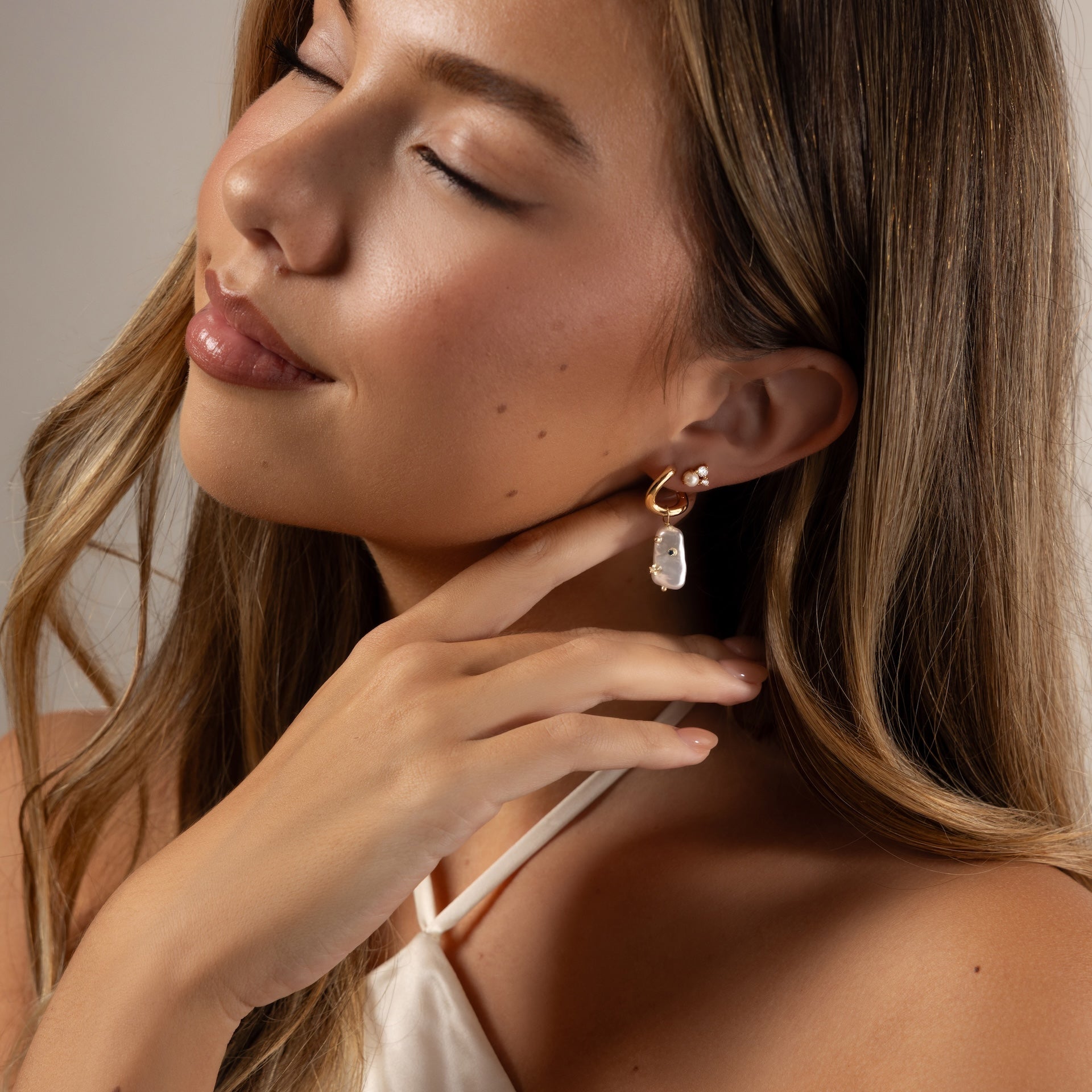A woman with long hair gently touches her Pearl Diamond Cluster Studs and smiles softly with her eyes closed.