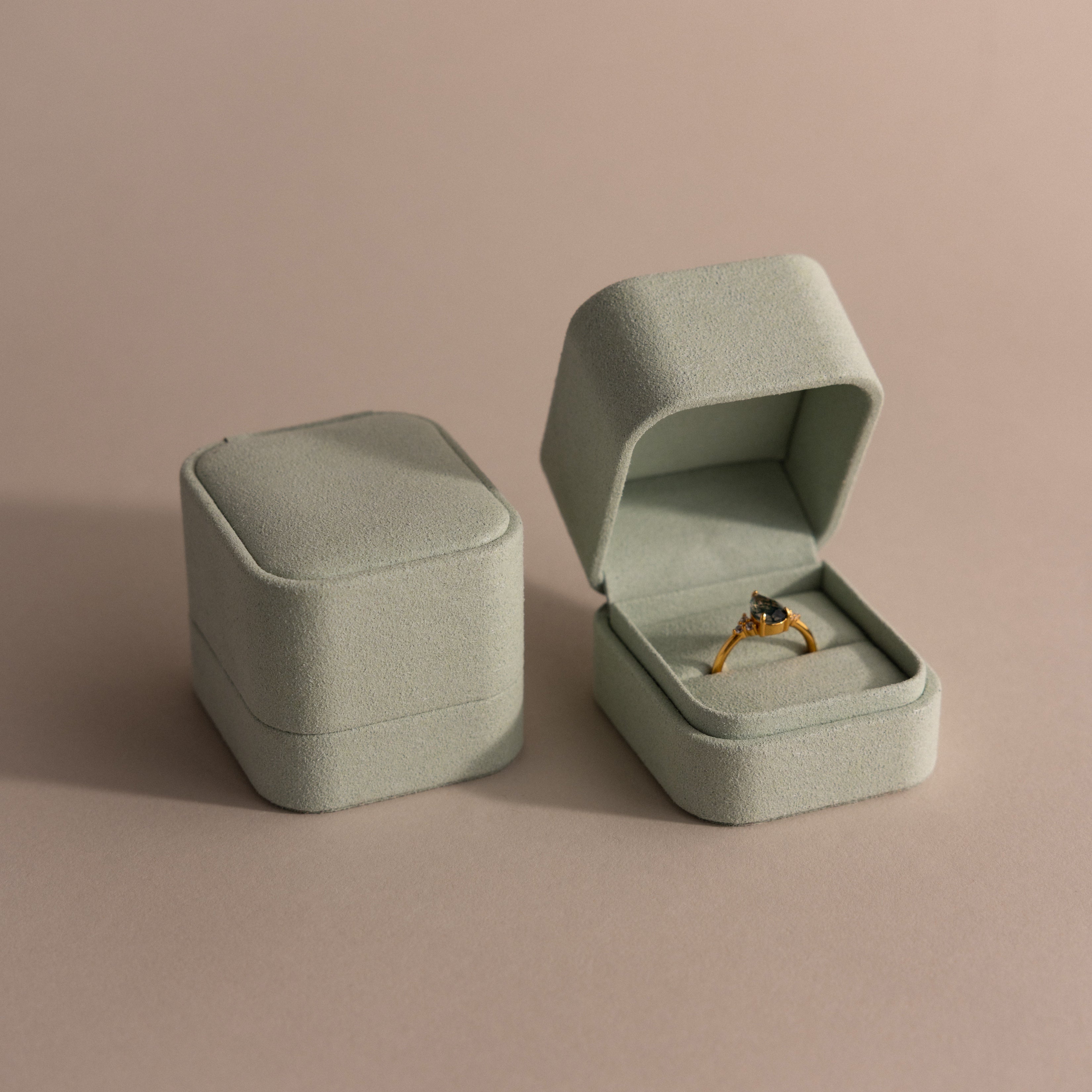 A gold ring with a dark stone is displayed in an open Green Suede Ring Box, next to a closed matching box, both set on a beige surface.