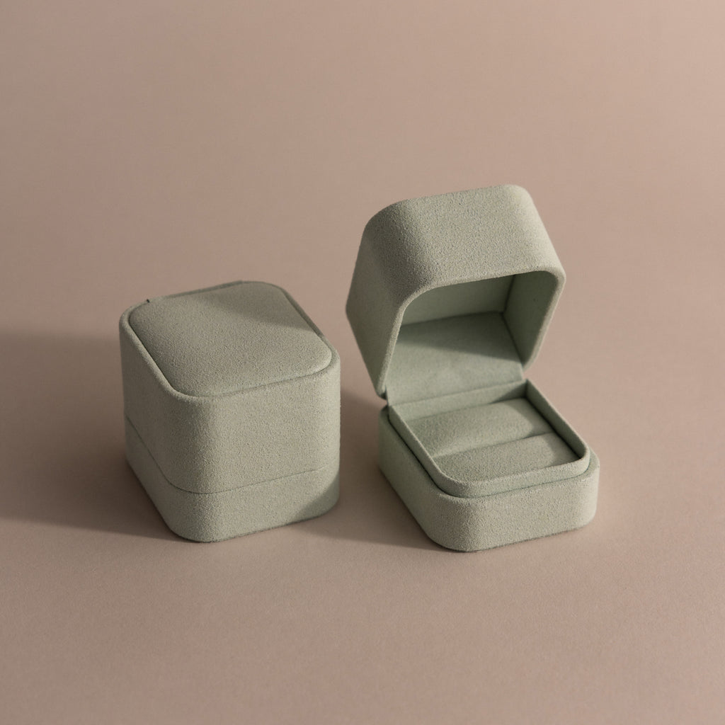 A closed and an open Green Suede Ring Box, featuring a minimalist design, rest on a neutral beige surface.