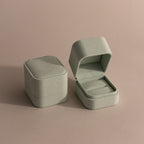 A closed and an open Green Suede Ring Box, featuring a minimalist design, rest on a neutral beige surface.
