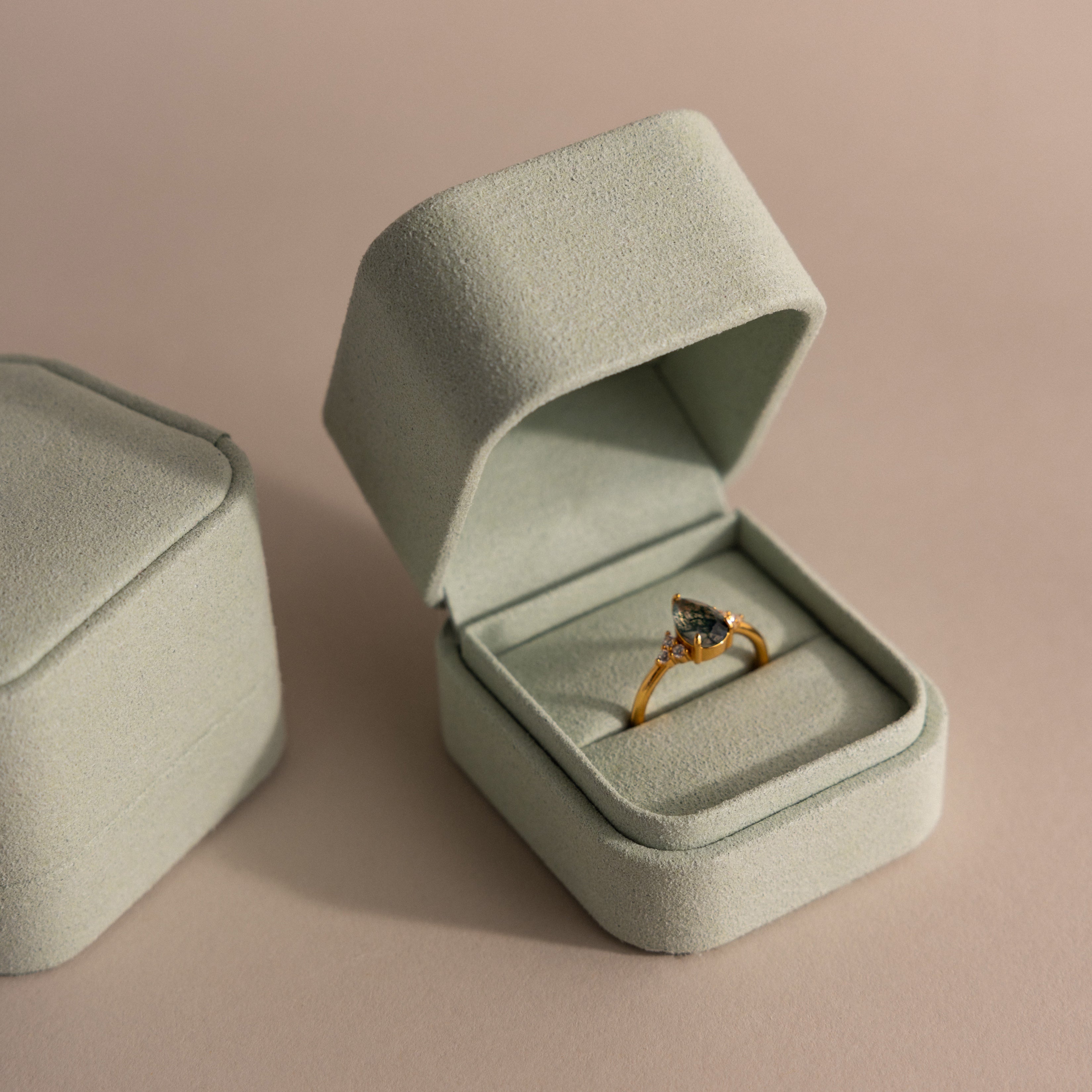 A Green Suede Ring Box with a minimalist design sits open on a soft beige surface, showcasing a gold ring set with a dark gemstone.