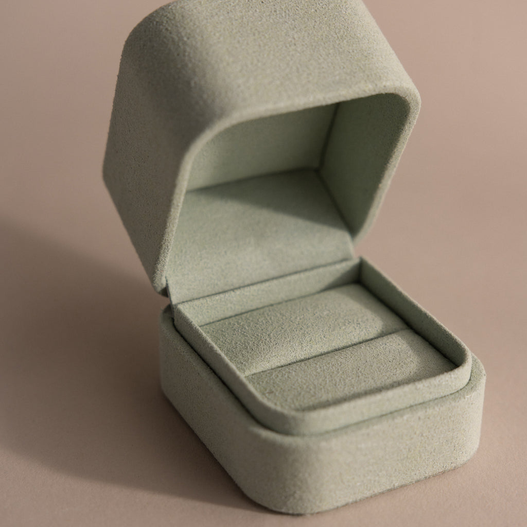 The Green Suede Ring Box features a soft, light green suede exterior and a minimalist design. Shown open on a beige surface.