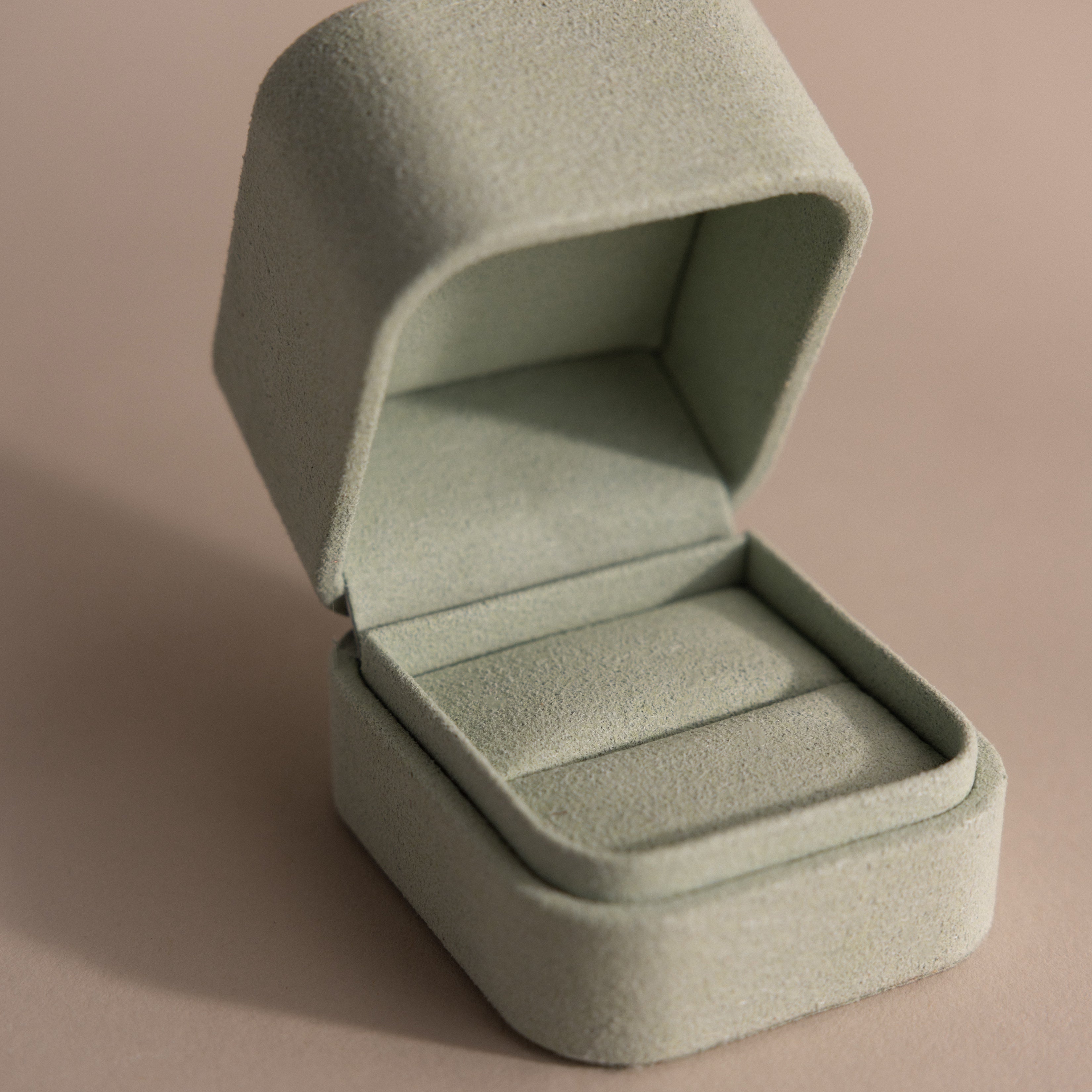 The Green Suede Ring Box features a soft, light green suede exterior and a minimalist design. Shown open on a beige surface.