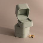 Two empty Green Suede Ring Boxes with a minimalist design are stacked, with a gold ring featuring a dark gemstone placed next to them.