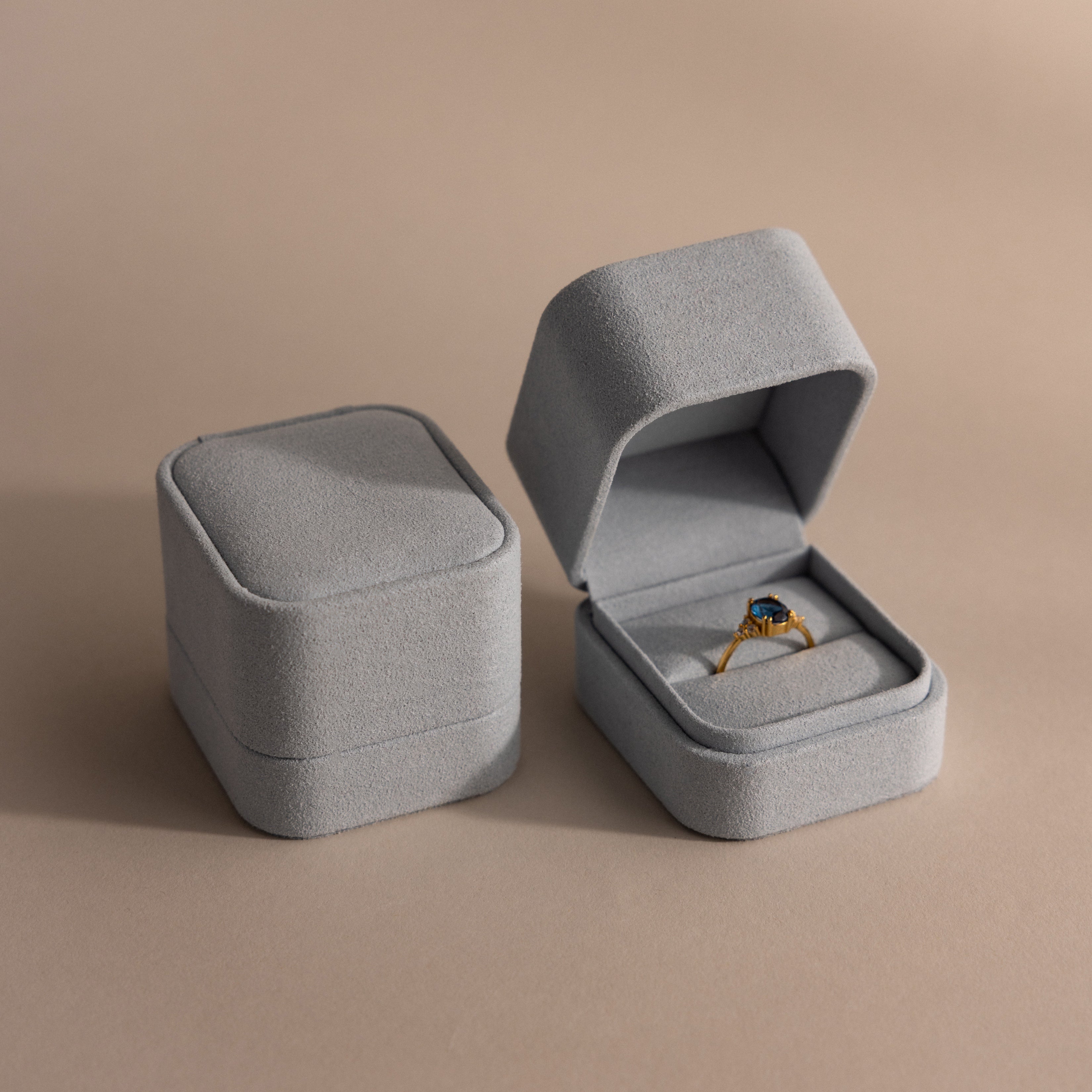 A Blue Suede Ring Box sits open on a beige minimalist surface, displaying a gold ring with a blue gemstone beside a closed jewelry box.
