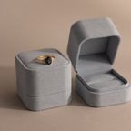 A gold ring with a dark gemstone rests in a Blue Suede Ring Box, placed next to an open empty matching box on a beige background for a minimalist look.
