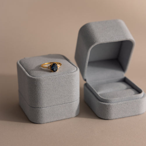 A gold ring with a dark gemstone rests in a Blue Suede Ring Box, placed next to an open empty matching box on a beige background for a minimalist look.