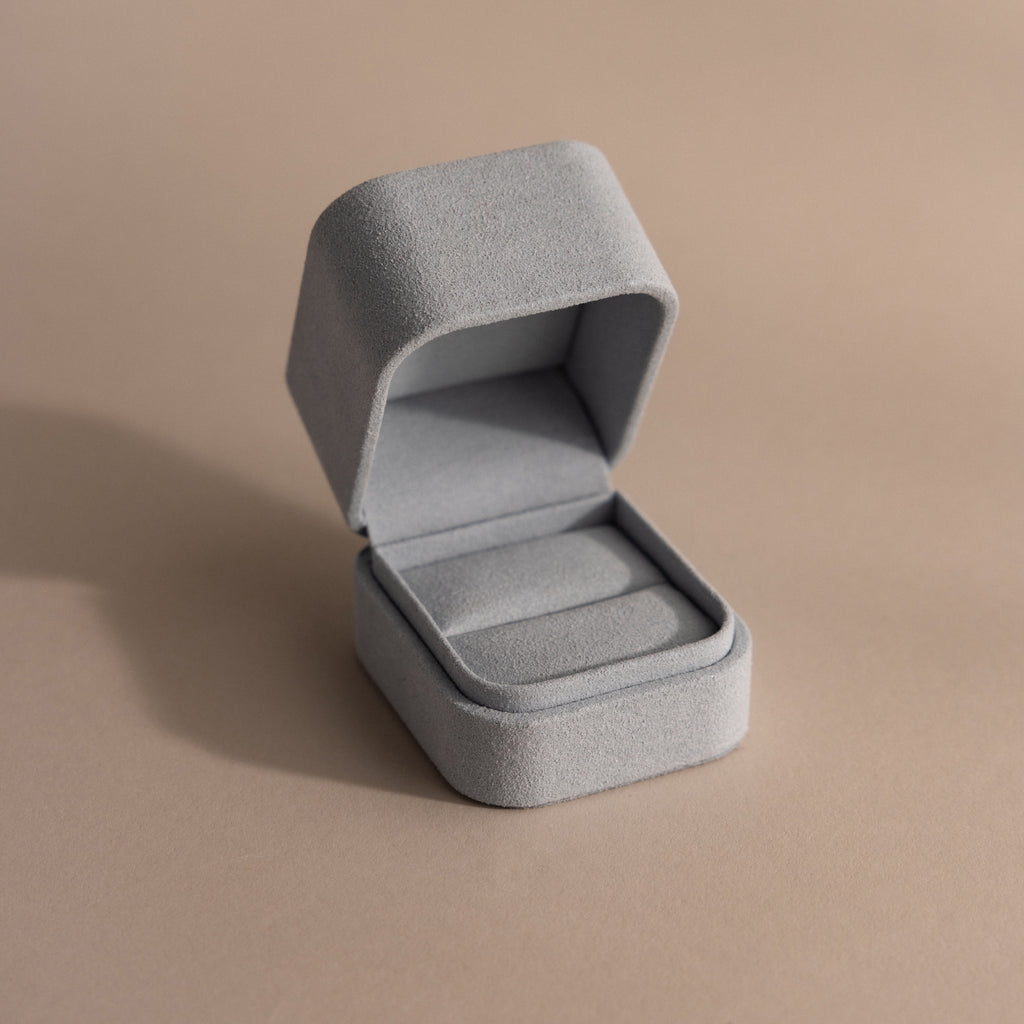 A blue suede ring box with a minimalist design sits empty on a neutral beige surface.