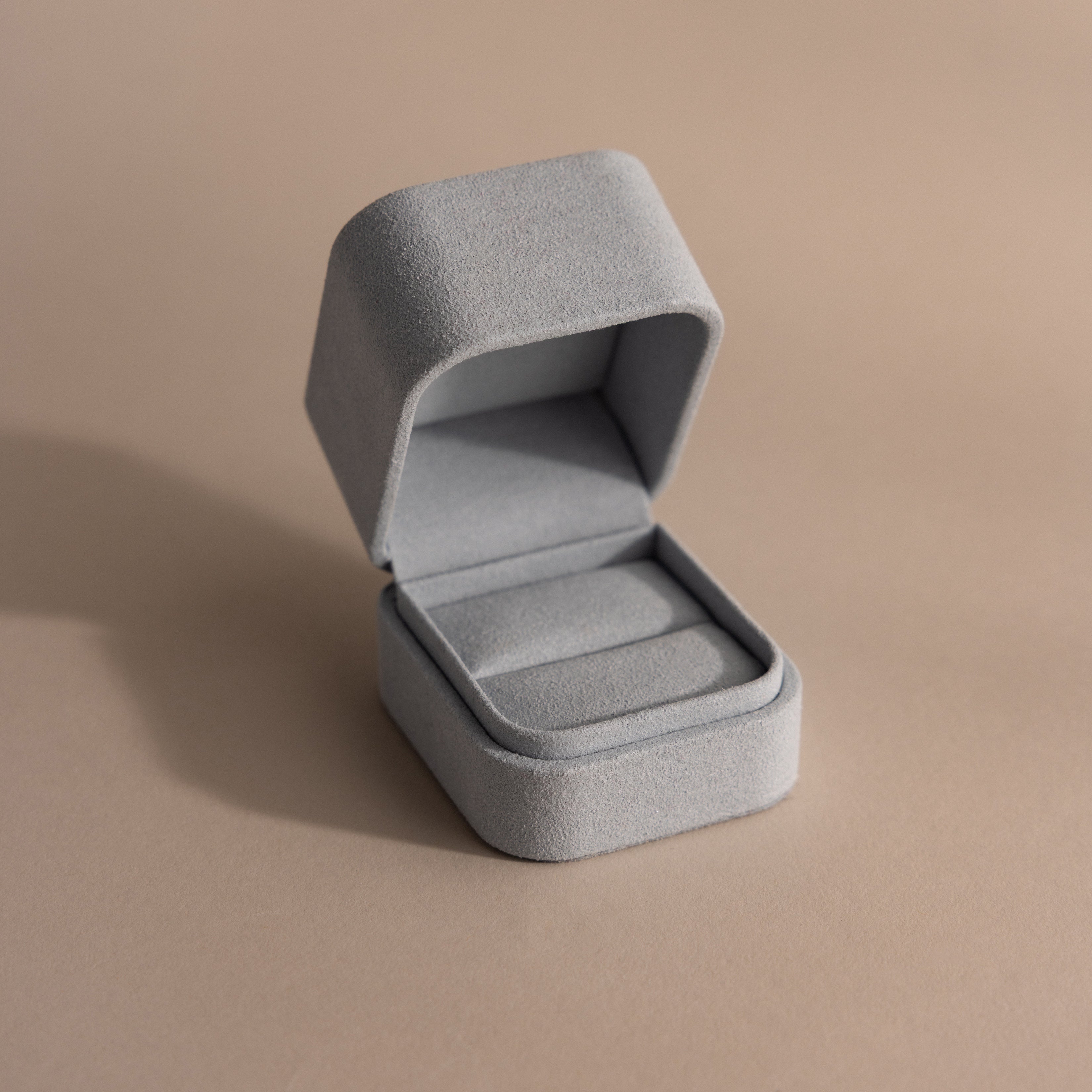A blue suede ring box with a minimalist design sits empty on a neutral beige surface.