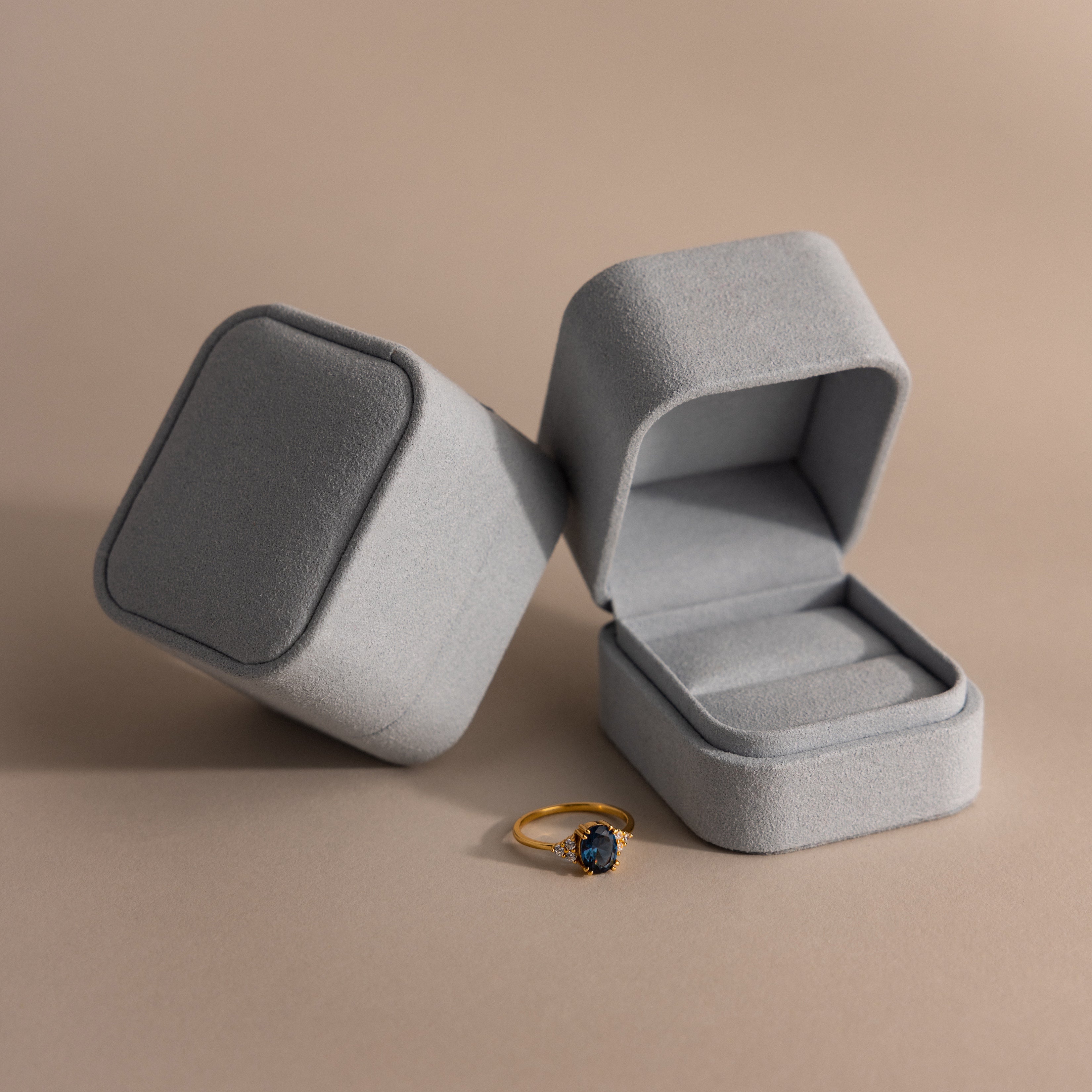A gold ring with a dark stone sits next to an open and closed Blue Suede Ring Box on a beige surface, highlighting minimalist design.