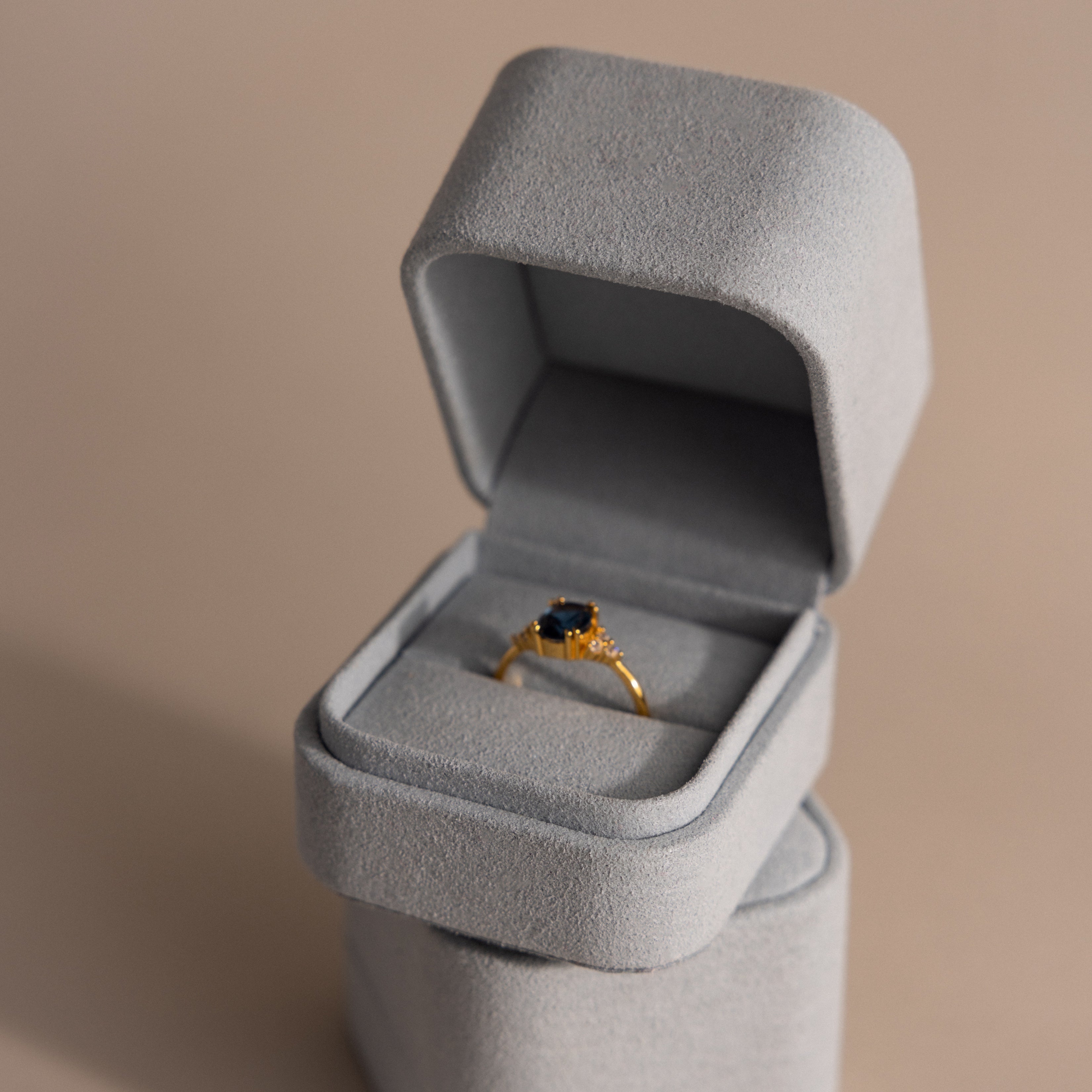 A gold ring with a dark gemstone displayed in a Blue Suede Ring Box on a beige surface, highlighting minimalist design.