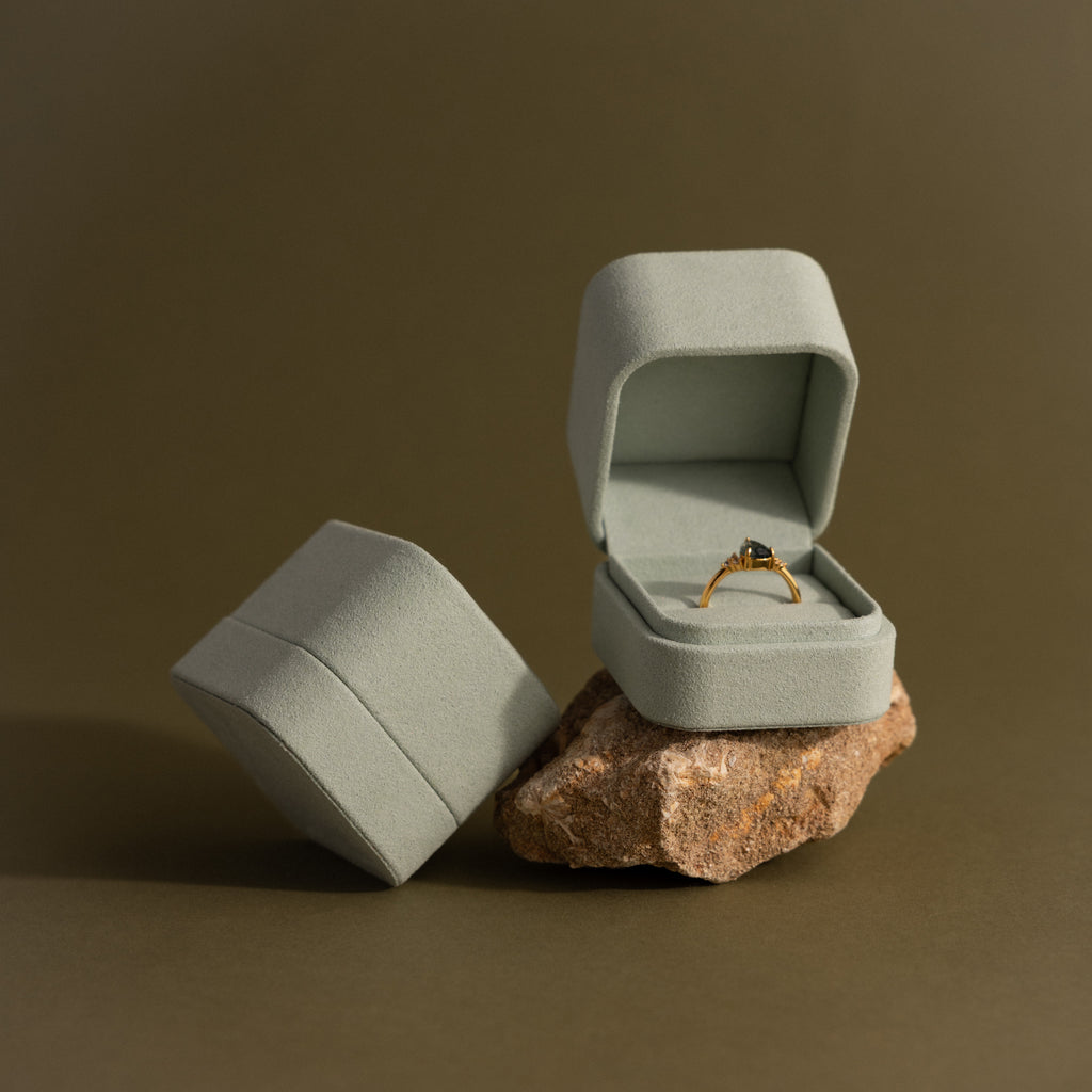A gold ring with a gemstone in a minimalist design is displayed in an open Green Suede Ring Box, set on a rock beside another box.