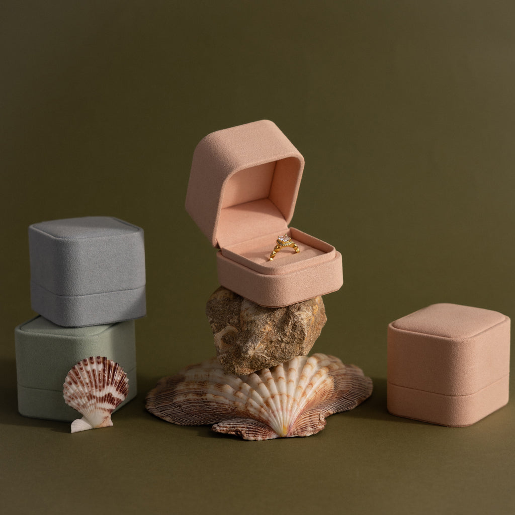 A Pink Suede Ring Box is displayed open on a rock, showcasing a gold ring alongside velvet jewelry boxes and seashells for a minimalist aesthetic.