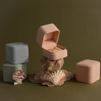 A Pink Suede Ring Box is displayed open on a rock, showcasing a gold ring alongside velvet jewelry boxes and seashells for a minimalist aesthetic.