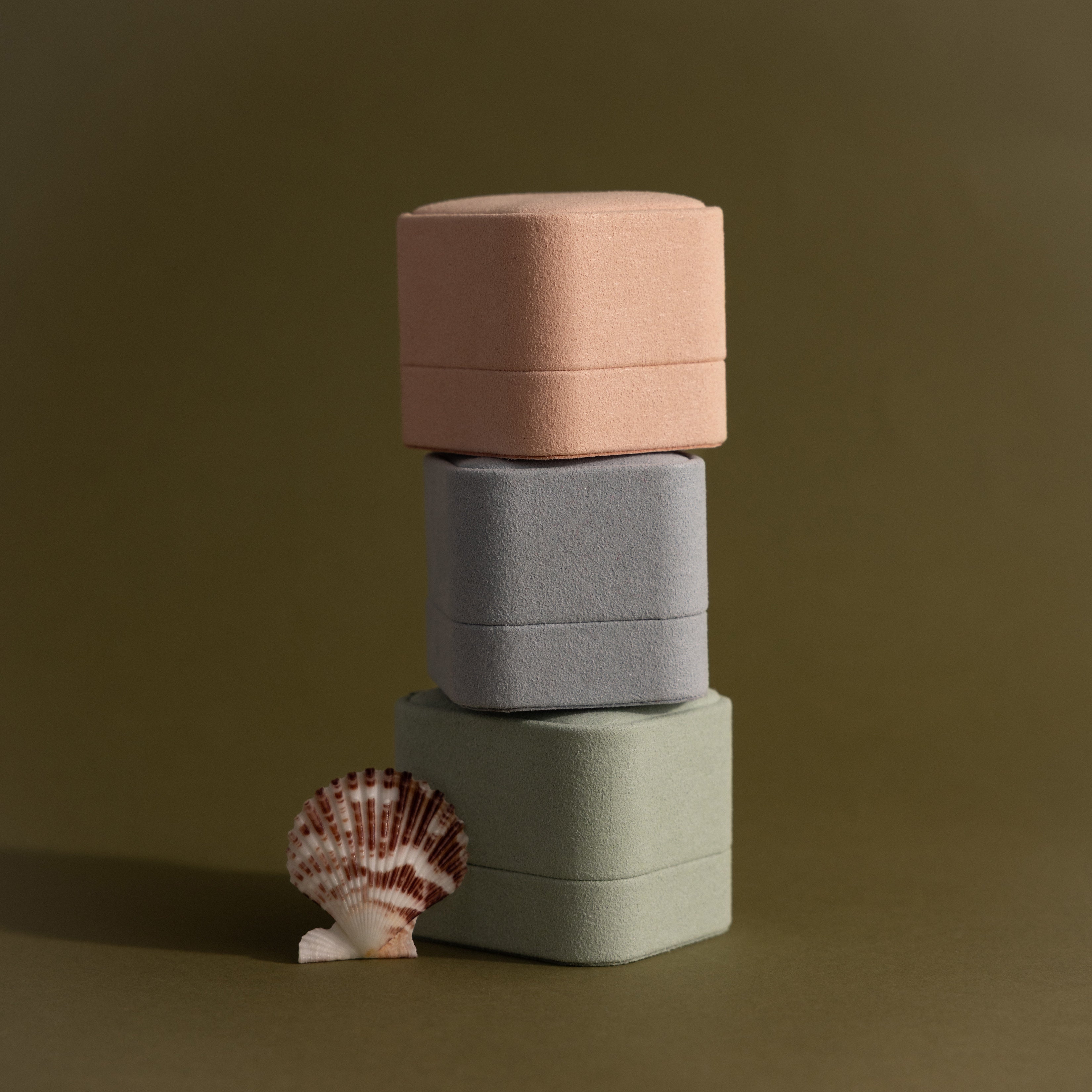 Three Suede Ring Boxes in pink, gray, and green are stacked beside a seashell on an olive background, highlighting their minimalist design.