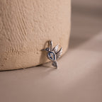 The Hummingbird Charm in Sterling Silver features a blue gemstone and delicate diamond accent, displayed on a textured beige surface with a soft background—an elegant and meaningful gift for any occasion.