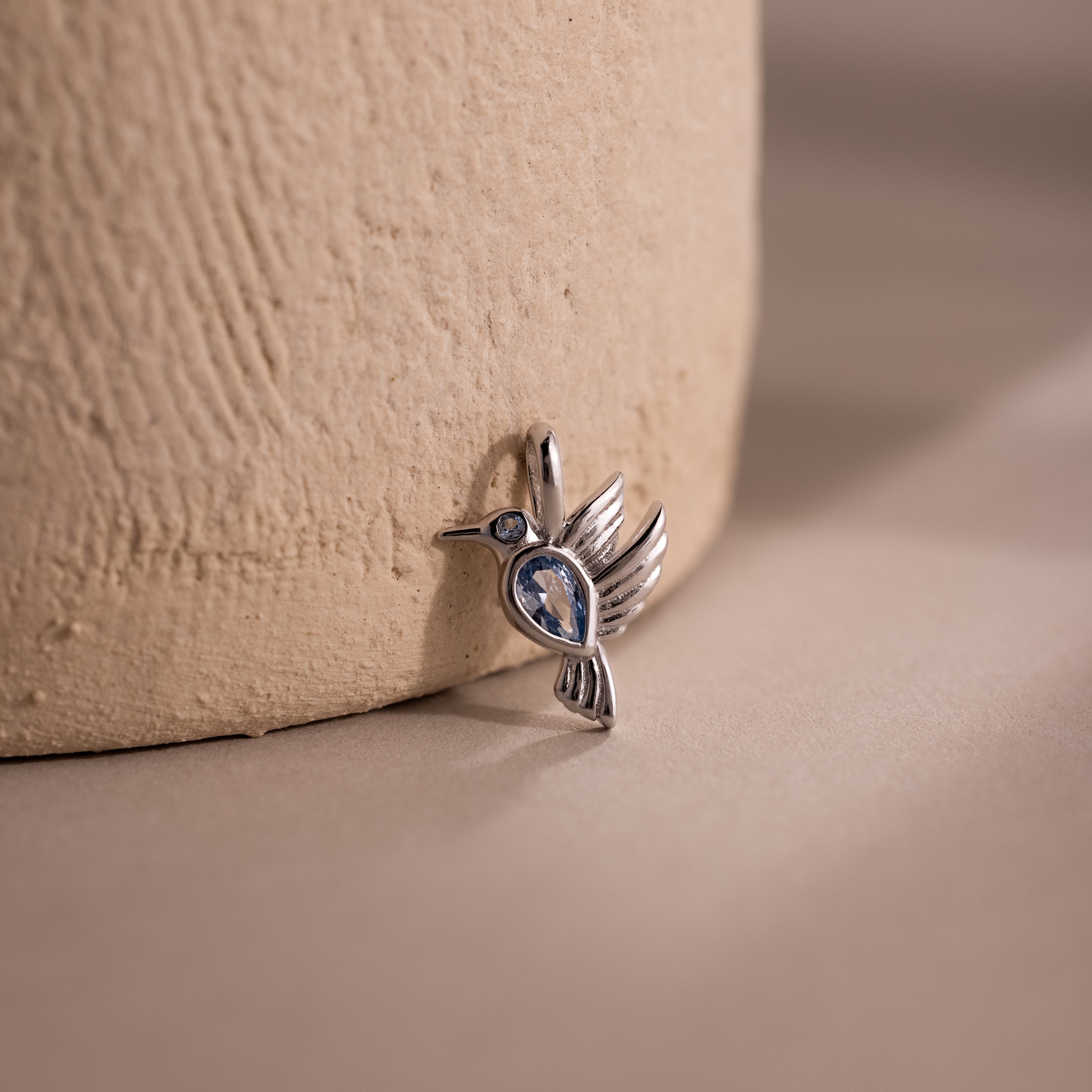 The Hummingbird Charm in Sterling Silver features a blue gemstone and delicate diamond accent, displayed on a textured beige surface with a soft background—an elegant and meaningful gift for any occasion.
