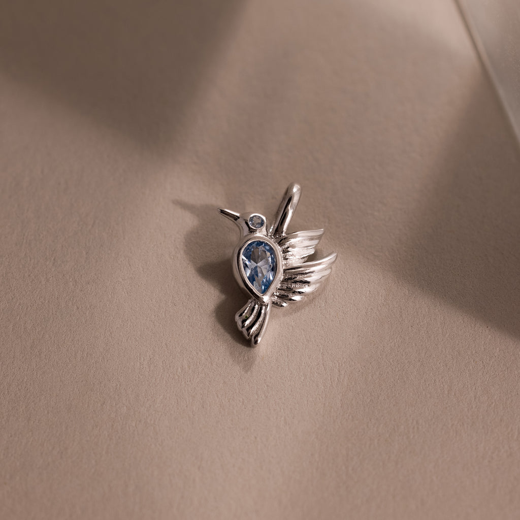 The Hummingbird Charm in Sterling Silver features a blue gemstone and diamond accent, displayed on a beige surface in gentle light—an elegant, meaningful gift.