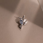 The Hummingbird Charm in Sterling Silver features a blue gemstone and diamond accent, displayed on a beige surface in gentle light—an elegant, meaningful gift.
