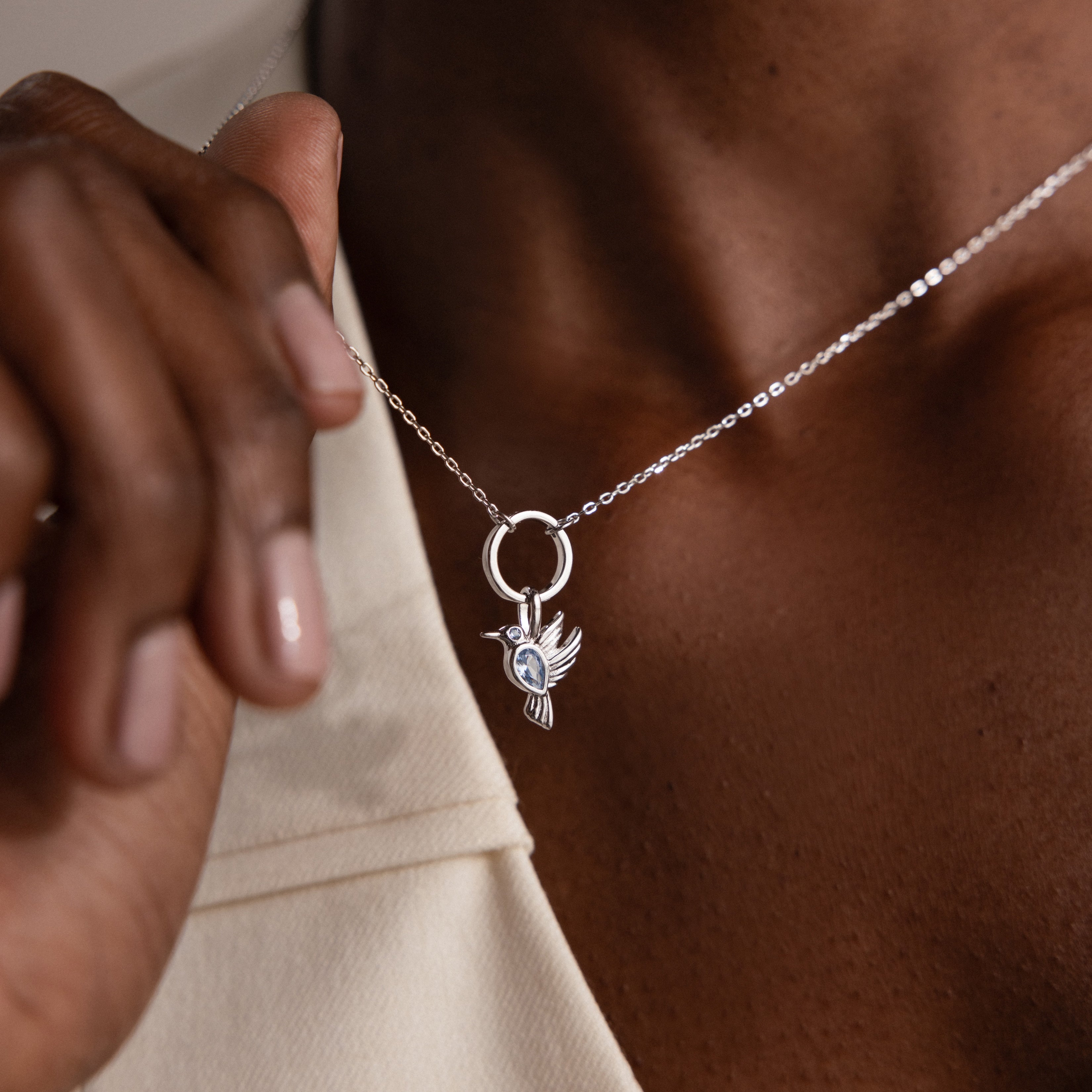 Wearing the Hummingbird Charm in Sterling Silver, someone holds the pendant near their collarbone—a beautiful and meaningful gift.
